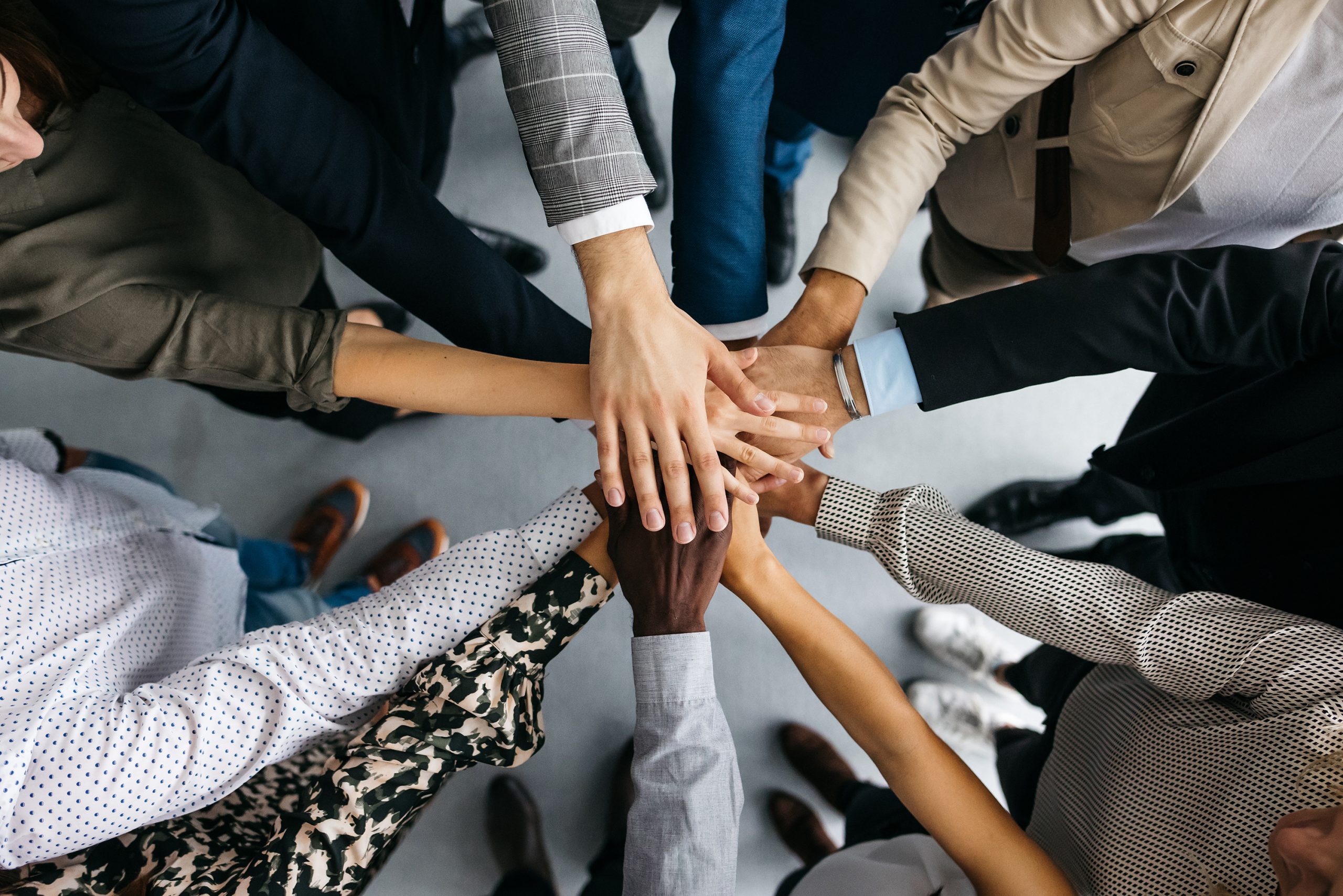 A high-angle shot of a group of male and female colleagues putting their hands together in an office. They are dressed in fashionable business clothes. Their faces are not visible, only their arms. Horizontal daylight indoor photo.